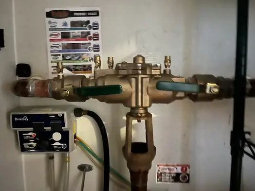 Brass backflow preventer on a restaurant water line beside a chemical dispenser