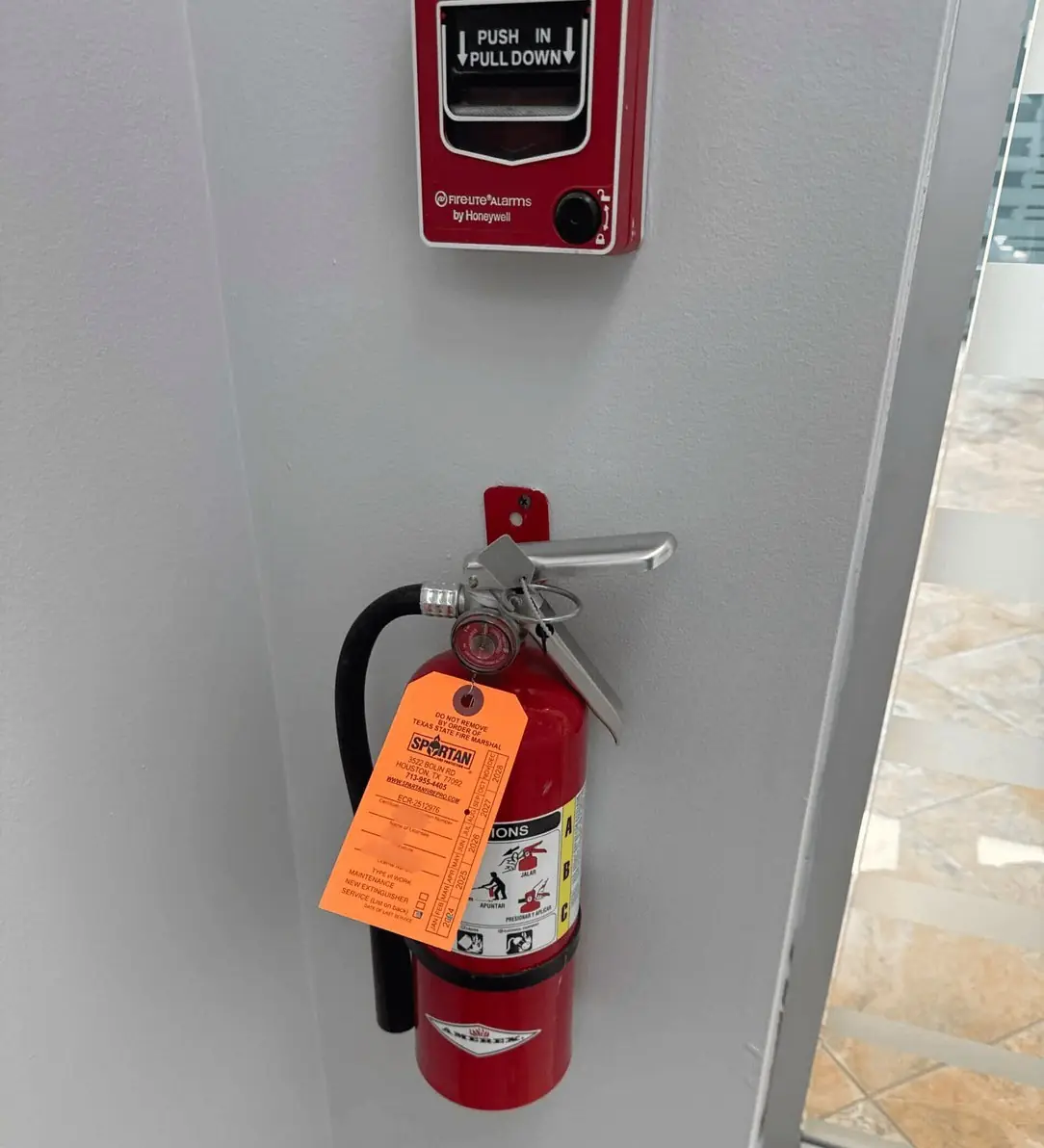Fire extinguisher with a Spartan inspection tag below a fire alarm pull station